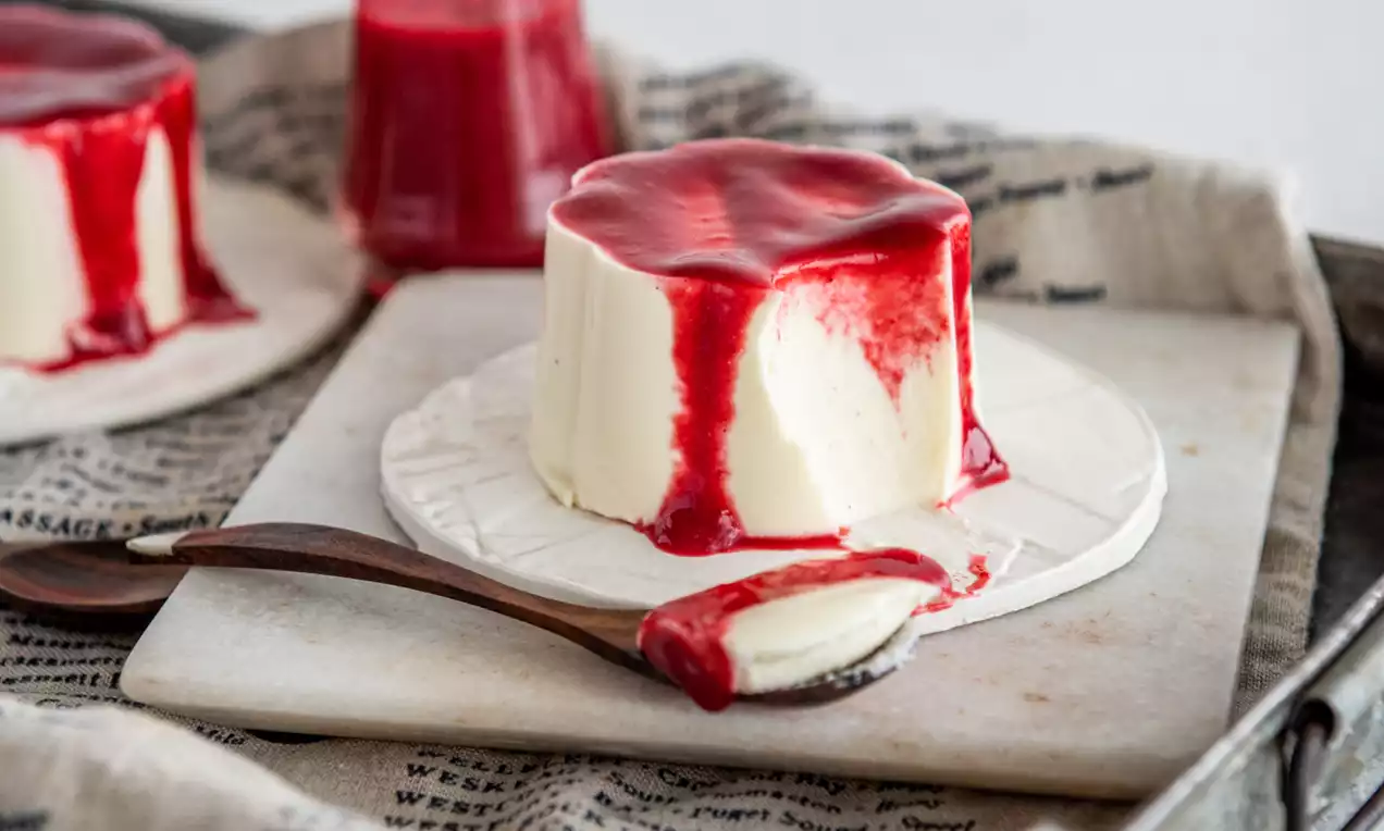 Picture - Vaniljefarvet panna cotta med glat overflade og skinnende rød frugtsauce, anrettet på en hvid tallerken med en ske på et metalfad