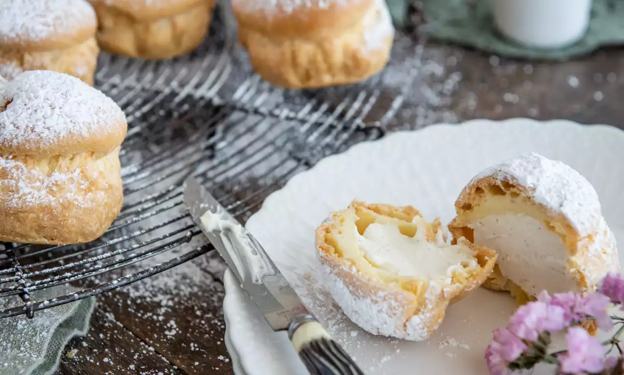 Picture - Lysebrune profiteroles med flormelis på en metalrist og hvid tallerken, flækket med cremet fyld, ved siden af lyserøde blomster og grønt stof