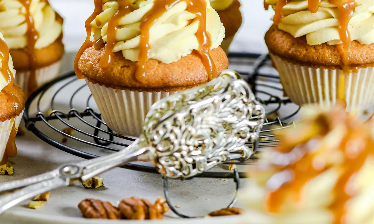 Picture - Gyldne cupcakes med hvirvler af lys smørcreme, karamelsauce og hakkede pekannødder på en rund bagerist med en ornamenteret ske